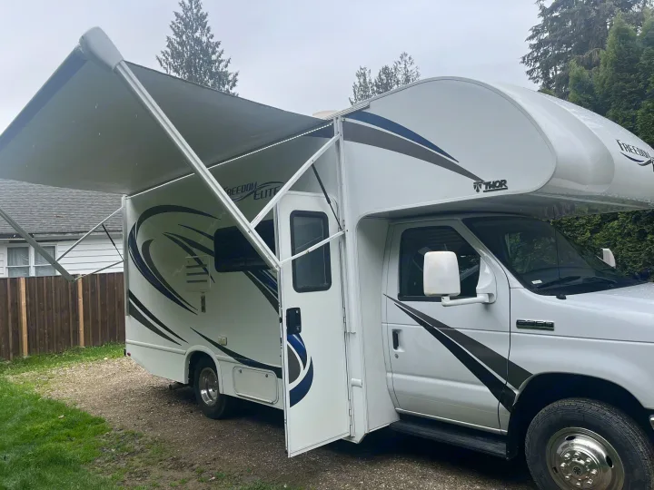 This white Thor Freedom Elite Class C RV rental is shown with its large awning extended, parked on a gravel drive.. Thor Freedom Elite 2019
