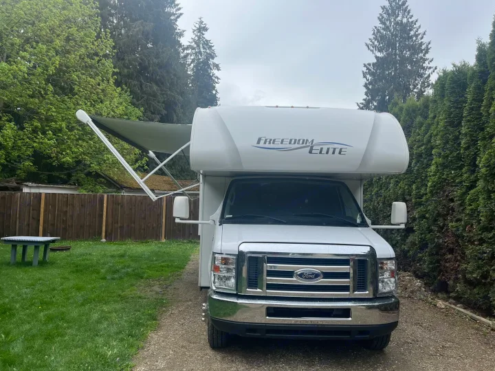 White "Freedom Elite" RV rental parked on a gravel driveway with an awning extended, surrounded by trees and grass.. Thor Freedom Elite 2019