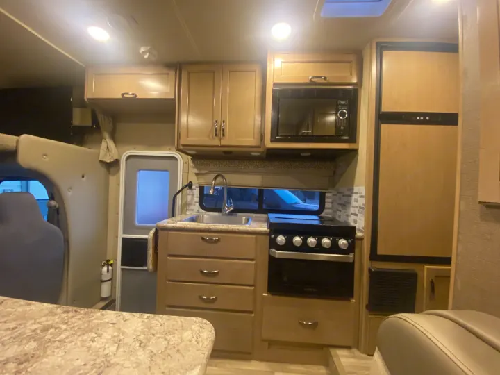 Interior of an RV rental: a modern kitchen with light wood cabinetry, microwave, oven, refrigerator, sink, and a fire extinguisher.. Thor Freedom Elite 2019