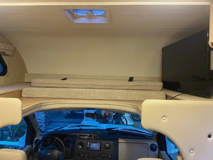 Inside an RV rental: the cozy cab-over bunk, dashboard, TV, and the outside view from the windshield.. Thor Freedom Elite 2019