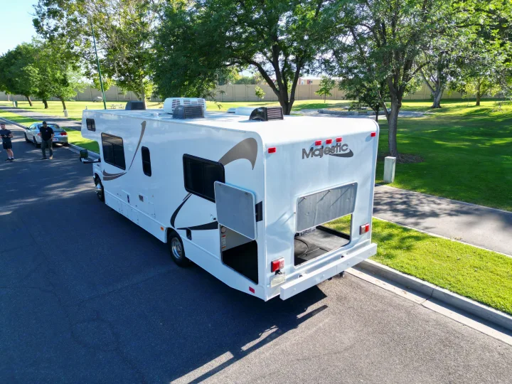 A white Majestic RV rental is parked with storage compartments open, two men and a car are nearby in a sunny park.. Thor Majestic 2019