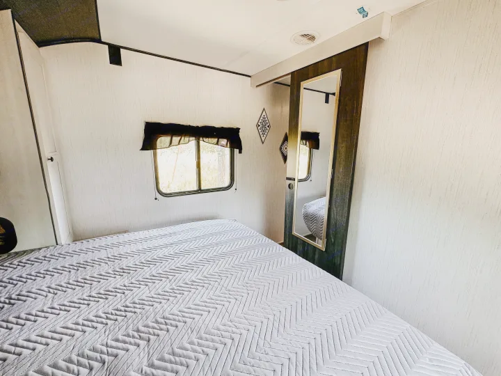 Cozy RV rental bedroom with a chevron-patterned bed, window, and full-length mirror for comfortable travel.. Heartland RVs Trail Runner 2022