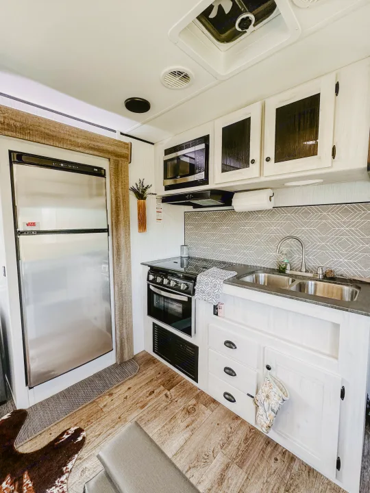 This RV rental kitchen boasts a refrigerator, microwave, stove, and double sink. Ample white cabinets, wood trim, and a patterned backsplash.. Heartland RVs Trail Runner 2022