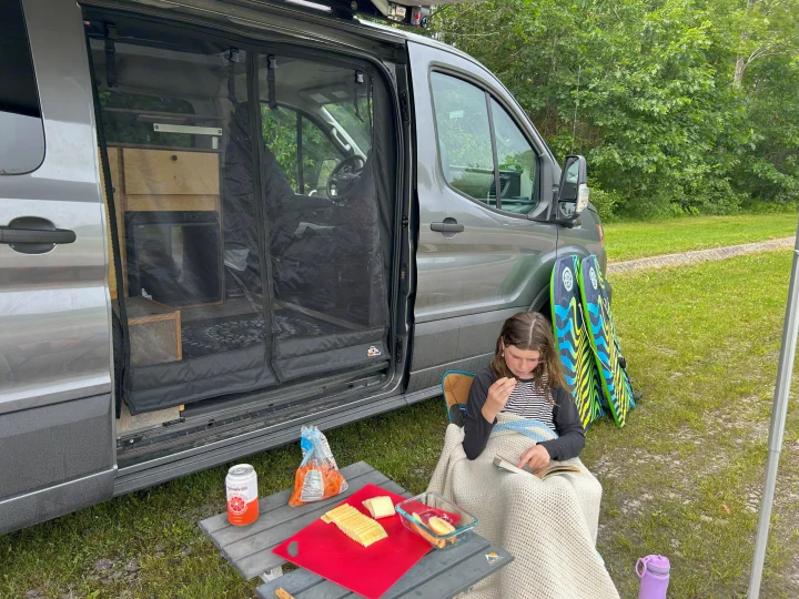 Child reads by a grey rv rental van, with snacks on a table and boogie boards outside for adventures.. Ford Custom Transit 2022