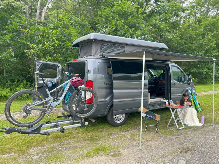 Pop-top RV rental with bikes on a hitch rack and an extended awning, offering a comfy outdoor camping setup for relaxation.. Ford Custom Transit 2022