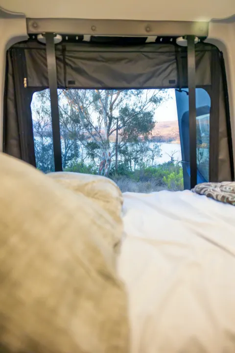 Cozy RV rental interior: a blurred bed leads to a clear view of a tranquil lake, trees, and distant hills.. Winnebago Revel 2023