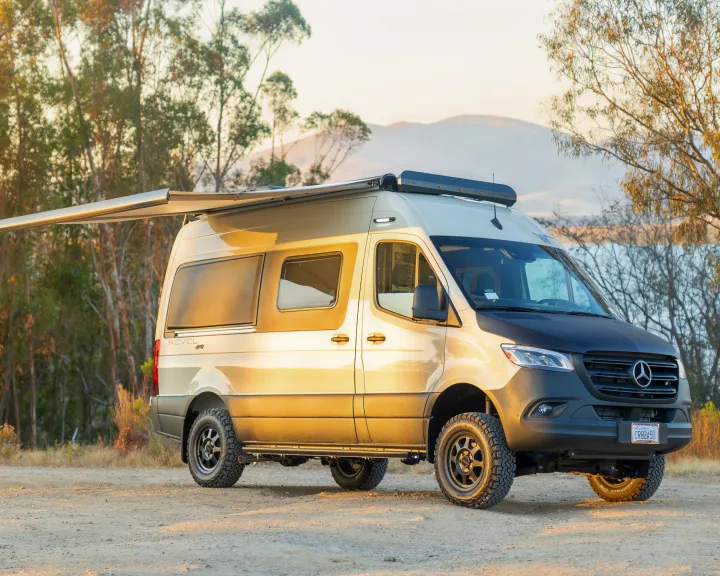 A tan Winnebago Revel 4x4, a rugged rv rental, parked with awning extended, ready for adventure by a lake.. Winnebago Revel 2023