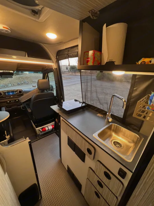 Inside a modern rv rental: kitchen with sink, induction cooktop, and driver's cabin. Ideal for compact mobile living.. Winnebago Revel 2023