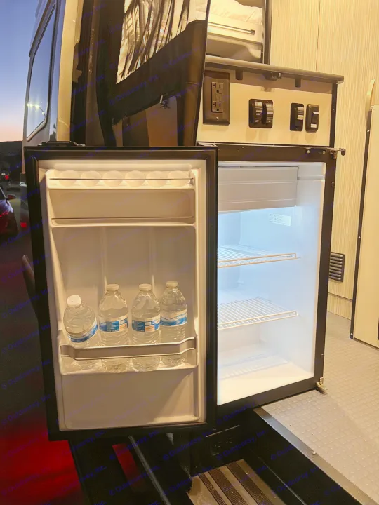 RV rental: Open mini-fridge with water bottles, empty freezer, control panel, & window view of dusk sky.. Winnebago Revel 2023