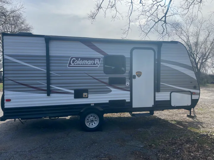 A gray and white Coleman travel trailer with its door visible, parked outdoors. This RV rental is ready for adventure.. Dutchmen Coleman Lantern LT 2019