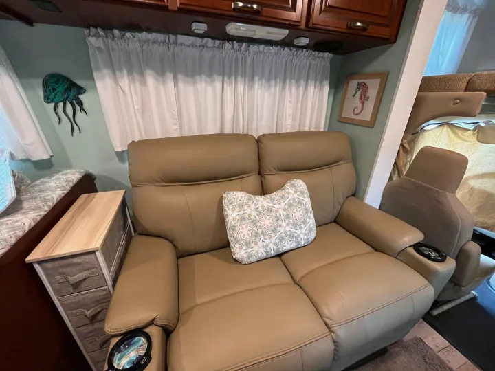 Relaxing RV rental living space with a comfy leather loveseat, decorative pillow, side table, and coastal wall art.. Thor Four Winds 2015
