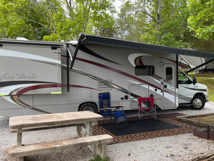 This RV rental showcases a comfortable campsite setup with an extended awning, outdoor chairs, and a concrete picnic table.. Thor Four Winds 2015