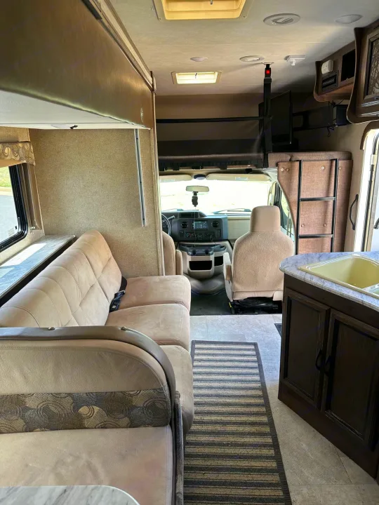 RV rental interior with a beige sofa, bunk bed above the driver's cab, kitchen counter with sink, and a striped rug on the floor.. Forest River Coachmen Freelander 2018