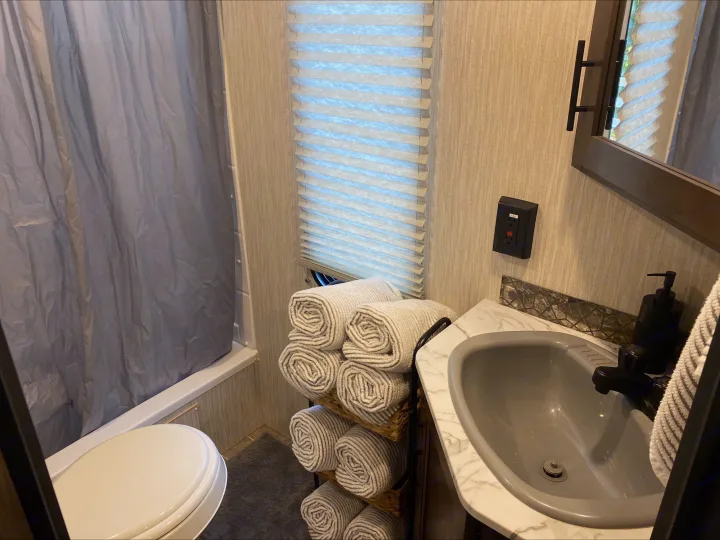 RV rental bathroom features a compact sink, toilet, shower, window with blinds, and neatly stacked towels.. Forest River Cherokee Grey Wolf 2021
