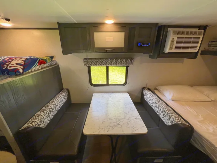 This RV rental interior shows a dining booth, bunk bed, and a main bed. Overhead cabinets and AC are also visible.. Aspen Trail 17BH 2021