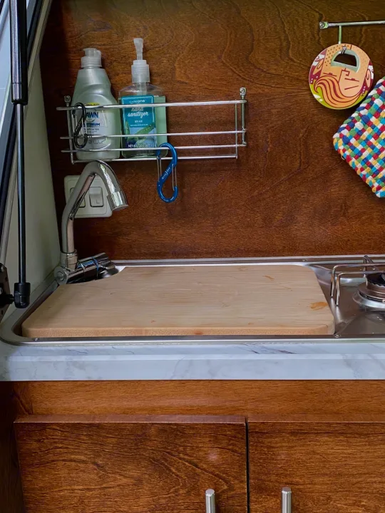 A compact RV rental kitchen area features a sink, faucet, cutting board, stove, and organized soap/sanitizer.. nuCamp T@G 2019