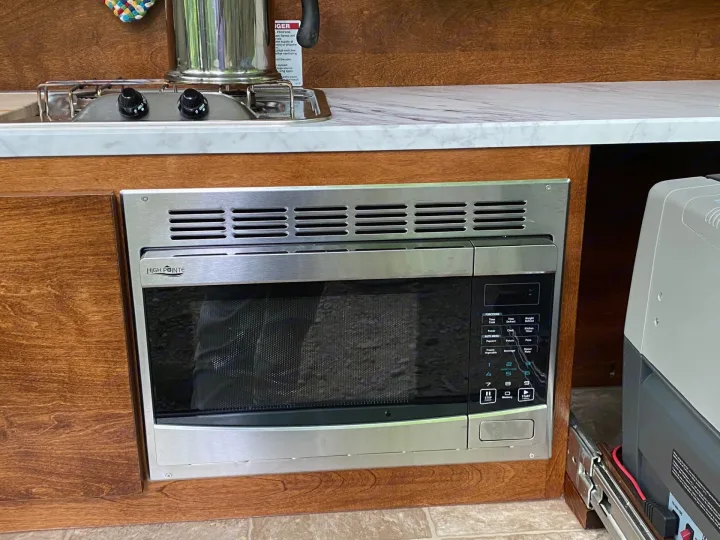 RV rental kitchen showing a built-in microwave, compact two-burner stovetop, and a portable fridge nearby.. nuCamp T@G 2019