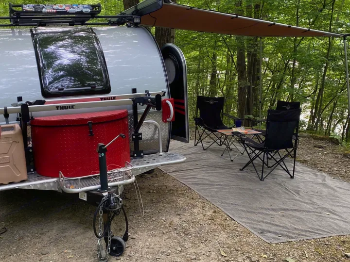 Cozy rv rental camping setup in a lush forest campsite, featuring a teardrop trailer with awning, chairs, and table for outdoor relaxation.. nuCamp T@G 2019