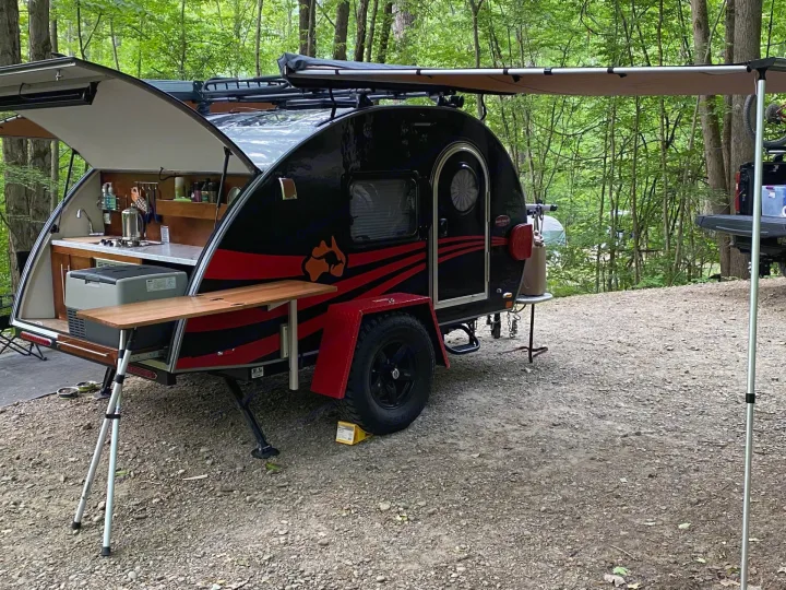 Adventure-ready RV rental: a black teardrop trailer with an open galley kitchen and awning, set up in a lush forest campground.. nuCamp T@G 2019