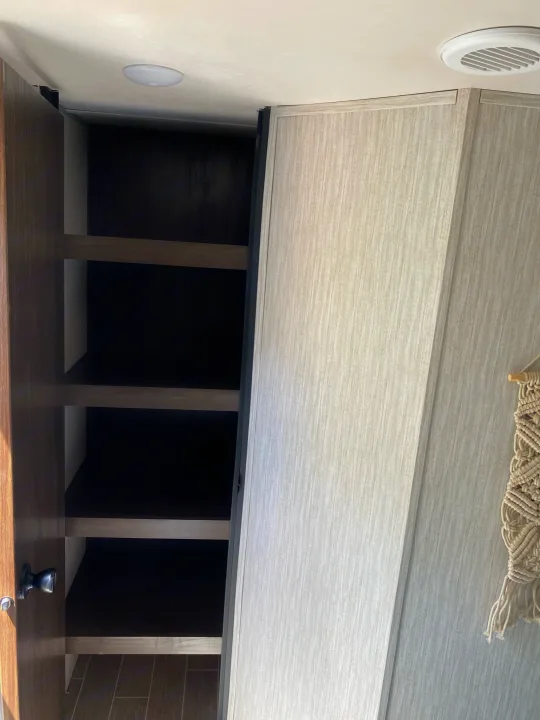 Empty, open pantry cabinet with shelves in an RV rental. Features bright ceiling light, vent, and macrame decor.. Forest River Cherokee Grey Wolf 2021