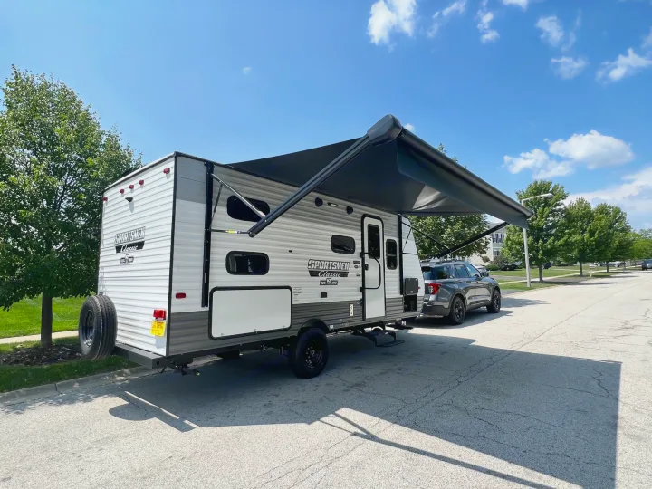 A Sportsmen Classic RV rental with its awning extended, parked on a sunny street next to a dark SUV.. KZ Sportsmen 191BHK 2022