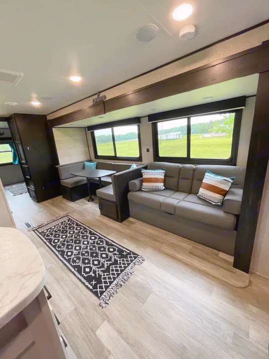Inside a bright RV rental: a modern living space with a grey sofa, cozy dinette, and big windows looking out to nature.. Jayco Jayflight 324BDS 2023
