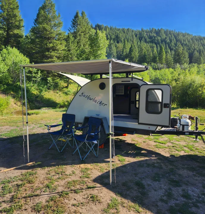 Bushwhacker RV rental set up in a scenic forest clearing with awning, chairs, and a dog, ready for outdoor adventure.. Braxton Creek Bushwacker 2023