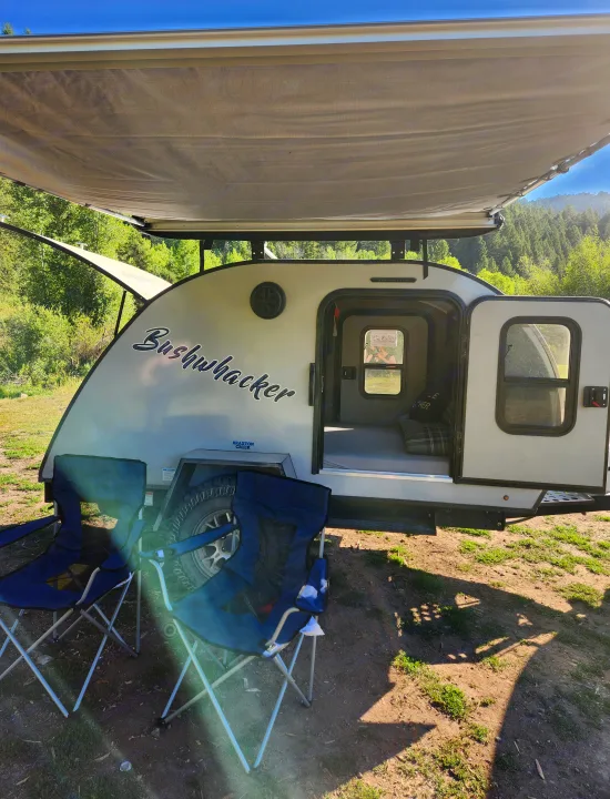 A white Bushwhacker teardrop RV rental with an extended awning and chairs set up for camping in a sunny mountain forest.. Braxton Creek Bushwacker 2023