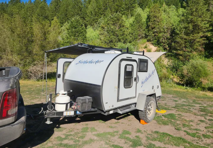 White Bushwhacker teardrop RV rental camper with awning extended in a scenic forest campground. Truck visible.. Braxton Creek Bushwacker 2023