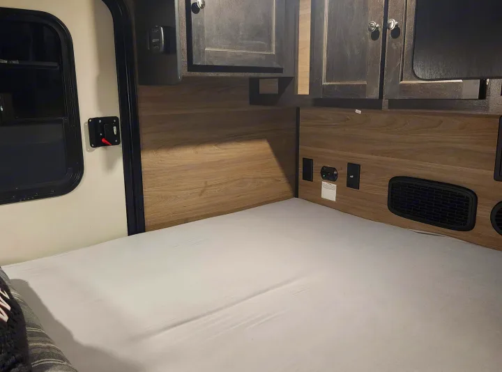 RV rental interior: cozy bed, wood walls with outlets/vents, and dark overhead cabinets by the door.. Braxton Creek Bushwacker 2023