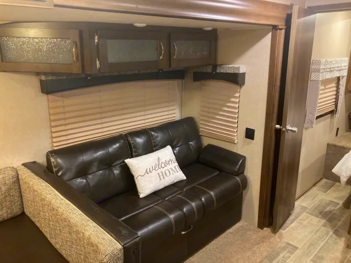Cozy RV rental interior featuring a dark leather sofa with a "welcome HOME" pillow, overhead cabinets, and window blinds.. Palomino Puma 31DBTS 2020
