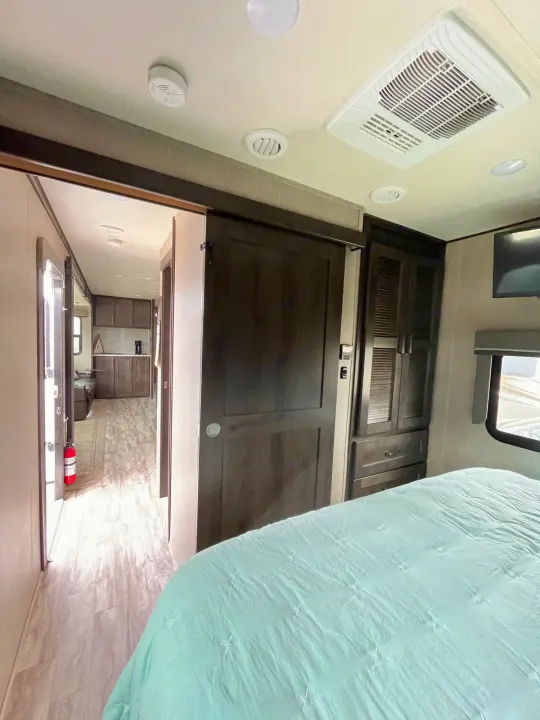 Inside an RV rental bedroom with a blue comforter, showing a sliding door leading to the kitchen and living area.. Grand Design Reflection 2022