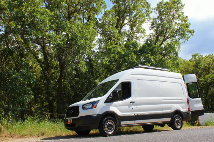 White camper van parked roadside by green trees. This RV rental is ready for scenic road trips and outdoor adventures.. Ford Transit 2017