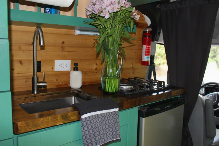 This RV rental's functional kitchen features a modern sink, stovetop, mini-fridge, wood walls, and vibrant teal cabinets.. Ford Transit 2017