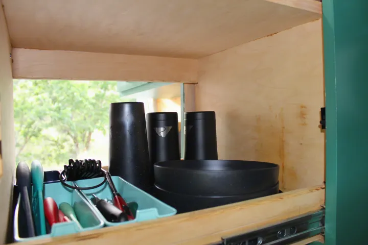 Efficient RV rental cabinet storage with black cups, bowls, and organized teal kitchen tools, showing a nature view outside.. Ford Transit 2017