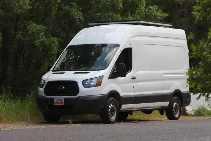 This white Ford Transit van with roof solar panels is ready for an RV rental adventure, parked on a road next to green trees.. Ford Transit 2017