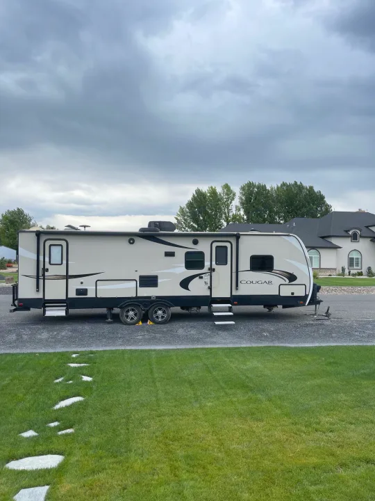 Modern Cougar travel trailer for RV rental, parked on a paved lot with a large house & green lawn under cloudy skies.. Keystone Cougar Half-Ton 2018
