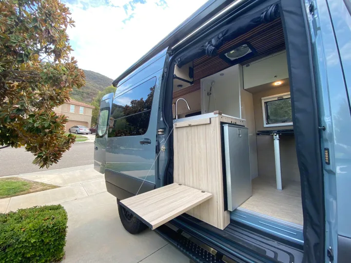 Gray RV rental camper van with open side door. Features mobile kitchen, fridge, and fold-out outdoor table. Perfect for adventures!. Mercedes Benz Sprinter 2500 2022