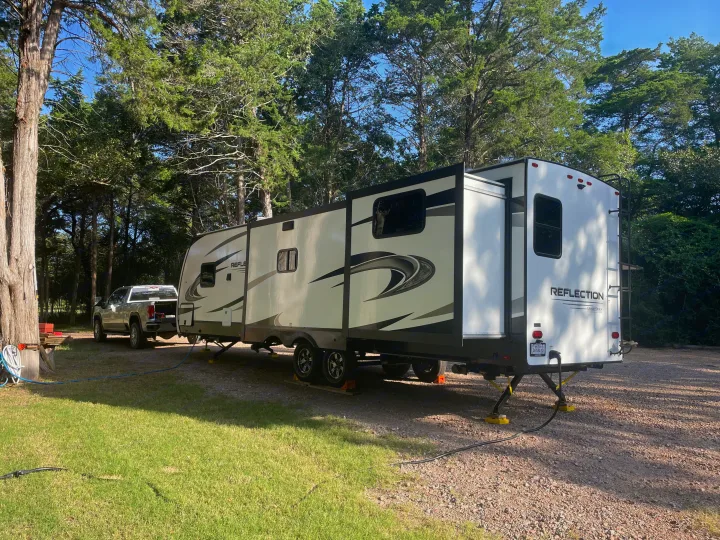 White RV rental & pickup truck parked at a tree-shaded campsite with hookups, ready for a comfortable outdoor stay.. Grand Design Reflection 2022