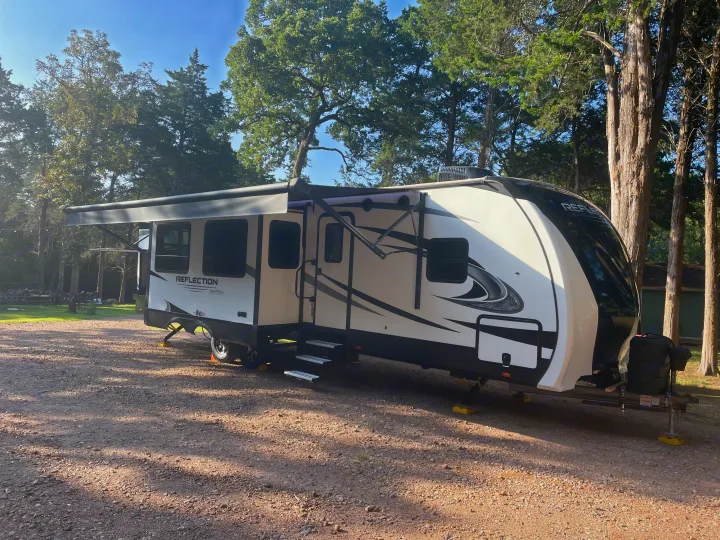 A white and black Reflection travel trailer, an RV rental, parked on a gravel campsite with awnings extended amidst green trees.. Grand Design Reflection 2022
