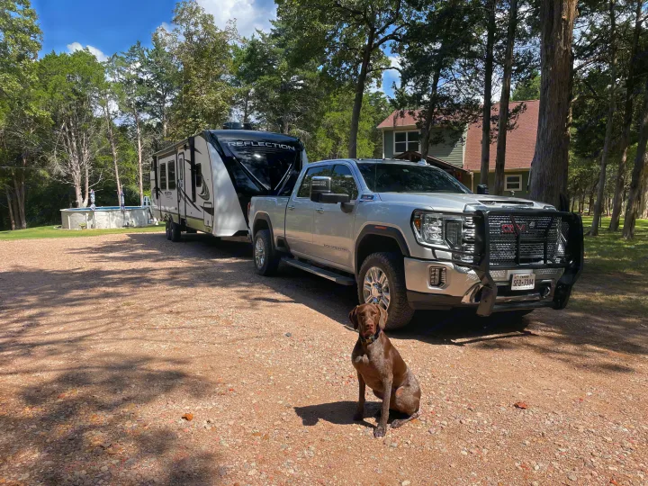 German Shorthaired Pointer sits by a silver GMC truck towing a Reflection travel trailer on a gravel drive. Perfect for your next rv rental.. Grand Design Reflection 2022