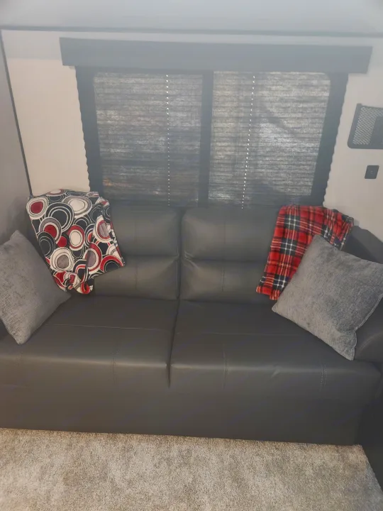 Inside a cozy RV rental: dark sectional sofa with plaid, patterned, and grey pillows, plus window blinds and carpet.. Keystone RV Springdale 2022