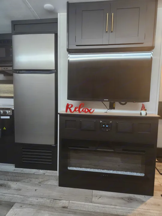 RV rental interior showing modern kitchen & living area with dark cabinets, Samsung TV, fridge, and electric fireplace.. Keystone RV Springdale 2022
