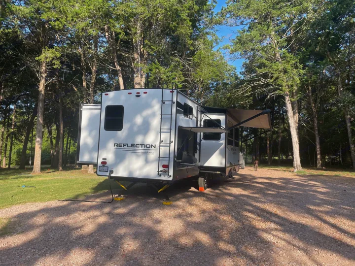 White RV rental parked on a gravel campsite under tall trees, with an extended awning. A dog rests nearby in the shade.. Grand Design Reflection 2022