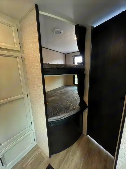 Inside an RV rental: a compact sleeping area with two neatly made bunk beds, privacy curtains, and light-colored cabinetry.. Jayco Jay Flight Baja Edition 2023