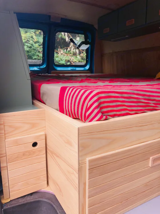 This RV rental features a cozy bed with a red striped blanket, custom wooden cabinetry, and practical storage.. Ford Econoline 2006