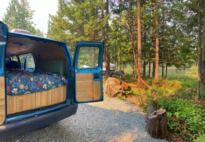 Inside a blue converted van (perfect RV rental) revealing a floral bed and wood storage, with a serene lake and forest campsite view.. Ford Econoline 2006