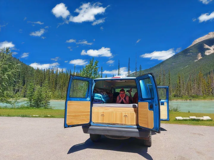 RV rental adventure: a blue campervan opens to a smiling person inside, overlooking a serene river, forest, and mountains.. Ford Econoline 2006