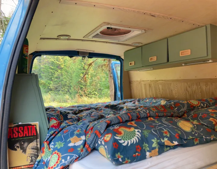 Cozy RV rental interior with vibrant bedding, green storage, window forest views, and a book.. Ford Econoline 2006