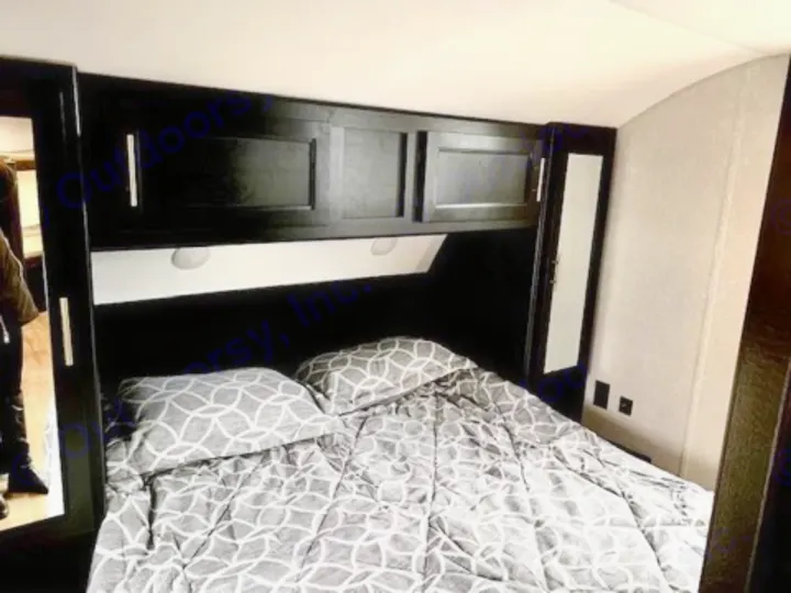 RV rental bedroom with gray patterned bed, dark overhead cabinets with lights, and a mirrored wardrobe for comfortable travel.. Forest River Cherokee Grey Wolf 2022